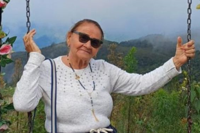 Imagem: Mulher sentada em um balanço ao ar livre, em um local cercado por vegetação e montanhas ao fundo sob neblina suave. Ela usa óculos escuros, blusa de manga longa clara com detalhes brilhantes e saia azul-marinho com listras claras. Traz uma bolsa azul a tiracolo. A mulher segura as correntes do balanço com as duas mãos e sorri levemente, em um cenário natural e tranquilo.