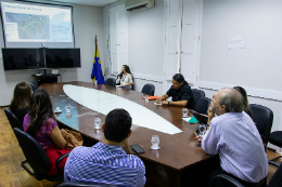 Imagem: foto de todos os participantes da reunião sentados ao redor de uma mesa e vendo apresentação do projeto numa projeção