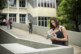 Imagem: Estudante encostada em uma parede próxima à biblioteca do Campus do Pici enquanto lê um livro (Foto: Viktor Braga/UFC)