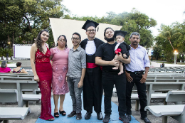 Imagem: foto de uma família em pé posando para a foto. Ao centro, um jovem de beca com faixa vermelha