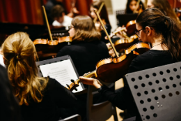 Imagem: foto de mulheres vistas de costas, sentadas, tocando violinos, com partituras a frente delas