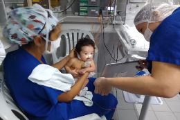 Imagem: foto de duas mulheres com roupas hospitalares segurando um bebê recém-nascido observando um tablet