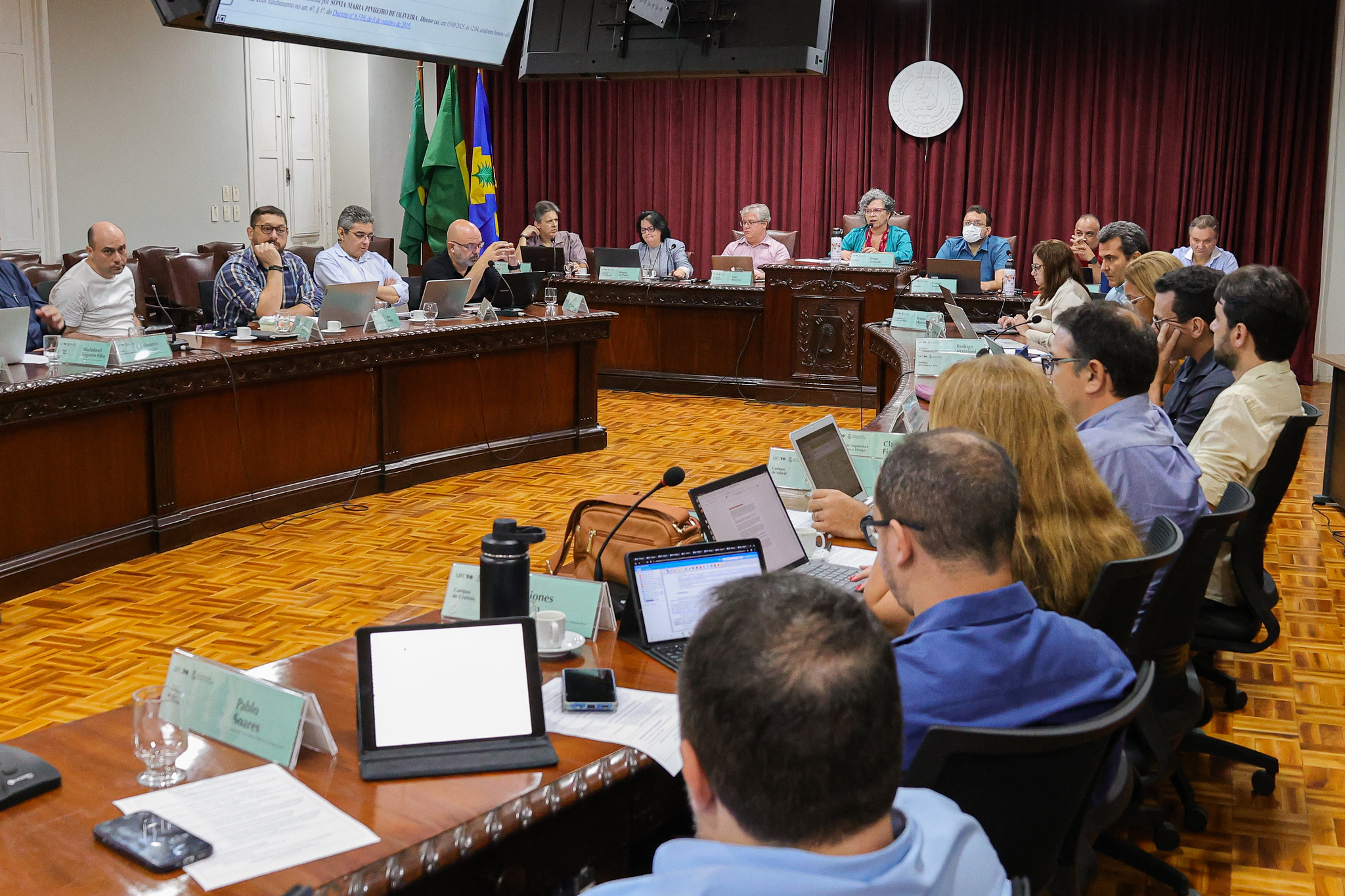Imagem: Reunião formal em uma sala ampla com cerca de vinte pessoas sentadas ao redor de uma mesa em formato de U. Os participantes estão atentos, muitos com laptops, papéis e tablets abertos à sua frente. A sala tem piso de madeira clara e cortinas vermelhas ao fundo, onde há um púlpito central elevado com uma mulher de cabelos grisalhos e roupas vermelhas, aparentando liderar a reunião. Bandeiras do Brasil e de um estado estão posicionadas no canto da sala. Um grande monitor suspenso exibe informações na parte superior da imagem. O ambiente transmite seriedade e concentração, típico de reuniões acadêmicas ou administrativas
