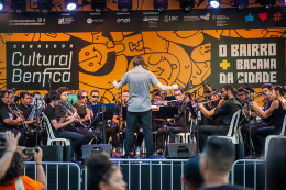 Foto do concerto da Banda Sinfônica da UFC-Fortaleza durante a programação do Corredor Cultural Benfica 2019. O maestro está de costas e rege o grupo no palco da Concha Acústica da UFC