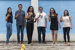Imagem: foto de cinco pessoas em pé, cada uma fazendo com as mãos uma letra em libras 
