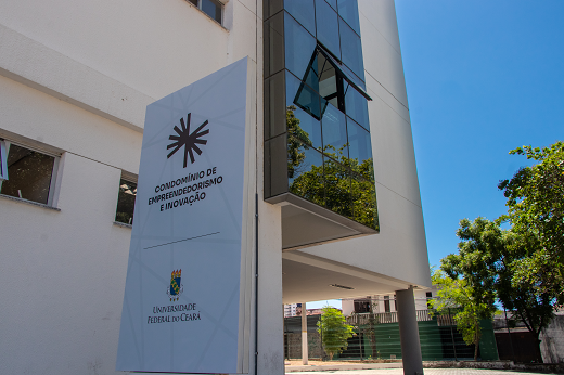 Imagem: Foto de parte da fachada do prédio do Condomínio do Empreendedorismo e Inovação. Em primeiro plano, a placa com a logo do Condomínio. Ao fundo, prédio branco (Foto: Viktor  Braga/UFC)