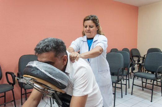 Imagem: Homem com blusa branca sentado em cadeira de massagem e mulher com bata branca fazendo massagem em sala com cadeiras e parede laranja