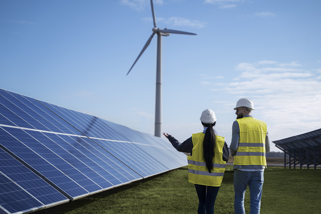 Imagem: Duas pessoas andando ao lado de painéis solares, tendo aerogeradores ao fundo (foto: freepik)