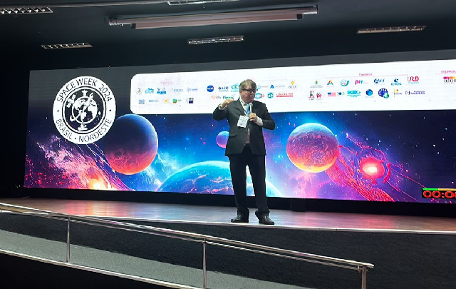 Imagem: Homem vestido de terno escuro fala em palco a frente de um banner com imagens espaciais onde se lê a frase Space Week 2024 (Foto: divulgação)  