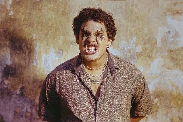 Mateus Fazeno Rock chamou atenção para o som produzido nas periferias de Fortaleza (Foto: Jorge Silvestre/Divulgação) Imagem: A imagem mostra um homem com expressão agressiva, gritando com os dentes à mostra e o rosto marcado por uma maquiagem escura ao redor dos olhos. Ele veste uma camisa simples e várias correntes douradas, posando diante de uma parede desgastada pelo tempo (Foto: Jorge Silvestre/Divulgação)