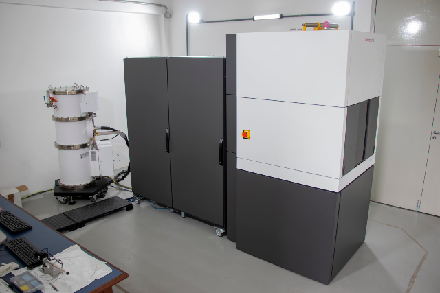 Grande equipamento de laboratório, do tamanho de um armário, com painéis fechados em tons de cinza e branco. Ao lado, há uma estrutura cilíndrica ligada por cabos, sugerindo um sistema científico de análise avançada. (Foto: Gutierrez Réges)