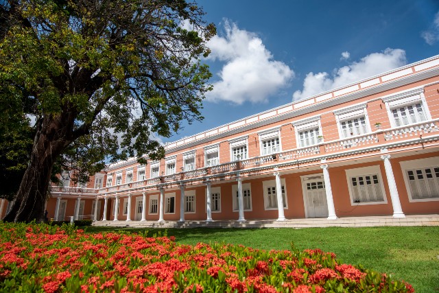 Foto do jardins da UFC. em primeiro plano pode-se ver grama verde e flores vermelhas. Ao fundo, um prédio rosa de dois andares (Foto: Victor Braga)