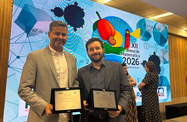 Foto mostra dois professores segurando certificados de premiação, em frente a um banner onde se pode ler: XII Bienal da Matemática (foto: Acervo pessoal) 