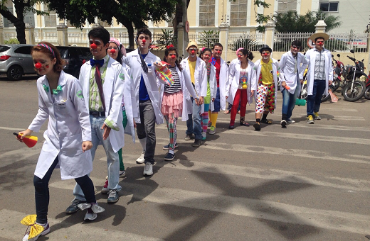 Imagem: foto de jovens com jaleco branco e adereços de palhaços atravessando uma rua