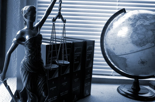 Imagem: foto em tom azul de uma estátua da balança que simboliza a justiça. Ao lado, um globo terrestre e, ao fundo, uma janela com persianas fechadas