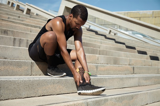 Imagem: foto de um jovem negro com roupa de esportes sentado em uma escadaria colocando as mãos no tornozelo