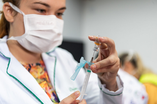 Foto de uma mulher vestida de jaleco e com máscara cirúrgica segurando uma ampola de vacina sedo furada por uma seringa