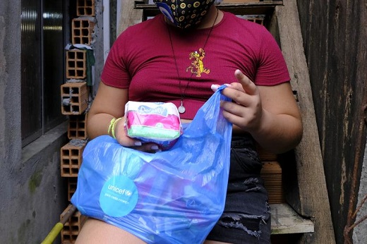 Imagem: Mulher, de máscara, segura pacote de itens de higiene menstrual (Foto: UNICEF/BRZ/Elias Costa)