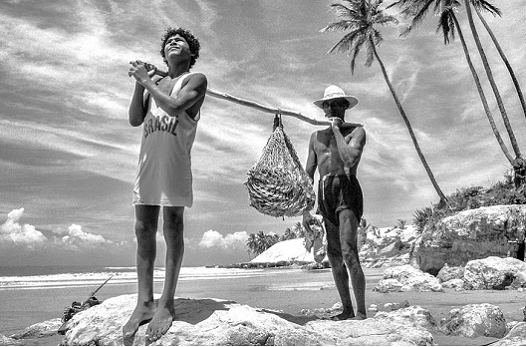 Imagem: Fotografia em preto e branco de Celso Oliveira, retratando um jovem e um homem em uma praia, segurando uma rede de pesca 