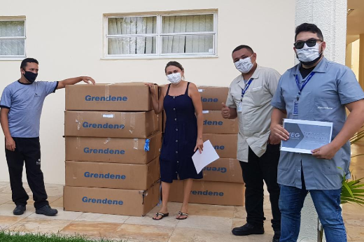 Imagem: Quatro pessoas em pé em uma sala, próximos a caixas contendo EPIs doados (Foto: Nelson Rodrigues de Souza/Grendene))