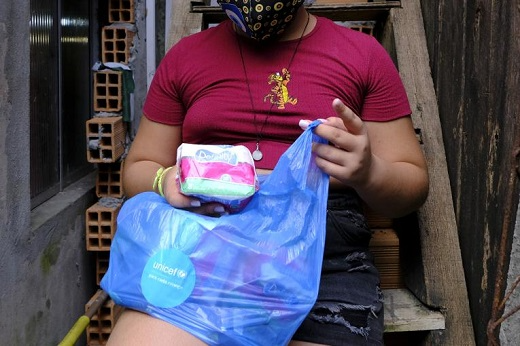 Imagem: foto de uma mulher negra sentada em um banco segurando uma sacola plástica azul e retirando dela um pacote de absorventes descartáveis