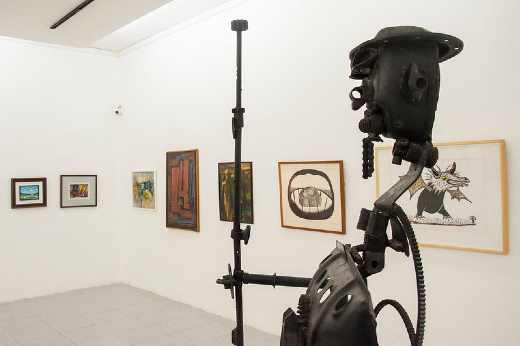 Imagem: escultura em ferro de um cangaceiro em primeiro plano, e pinturas ao fundo, em sala do MAUC 