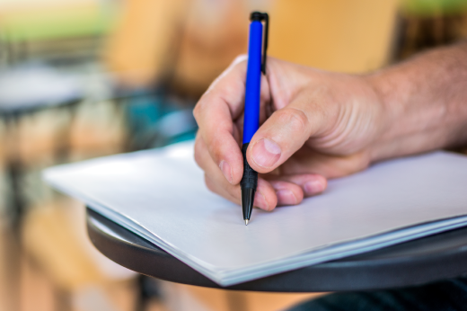 Imagem: foto da mão de uma pessoas segurando uma caneta e escrevendo num papel em cima de uma carteira