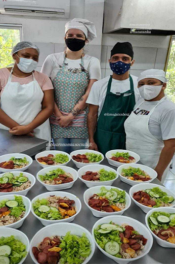 Imagem: Cozinheiros em frente a diversas marmitas expostas em mesa de cozinha industrial (Foto: Divulgação/EGA-UFC)