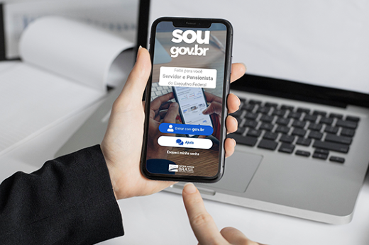 Imagem: foto focada nas mãos de uma pessoa segurando um aparelho de celular, cuja tela mostra o aplicativo SouGov