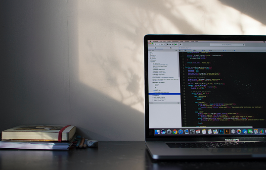 Imagem: caderno sobre uma mesa, ao lado de um notebook com a tela aberta, cheia de linguagem de computador. Os códigos estão todos coloridos (Imagem: Emile Perron/Unsplash)