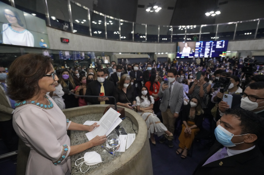 Imagem: Em primeiro plano, Izolda Cela com um vestido cinza, faz um discurso com papeis na mão em uma tribuna; ao fundo, multidão lota Plenário da Assembleia (Foto: Carlos Gibaja/Governo do Estado)