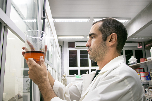 Imagem: Homem vestido de bata branca segura um frasco de laboratório com líquido laranja dentro em ambiente de laboratório.