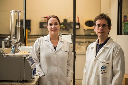 Foto: Pesquisadora do lado esquerdo, vestida de bata branca e pesquisador do lado direito vestido de bata branca em ambiente de laboratório.
