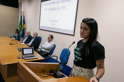 Imagem: A coordenadora da UFC Inova, Ana Carolina Matos fala em primeiro plano no púlpito do Auditório da Reitoria. (Foto: Viktor Braga/ UFC Informa)