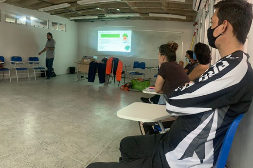 Imagem: Em primeiro plano, do lado direito, grupo está sentado, assistindo a uma aula. Do lado esquerdo ao fundo, vê-se professor de caça jeans e camisa cinza. Perto dele, no centro da imagem, um telão e vários equipamentos relacionados a poda (Foto: Divulgação)