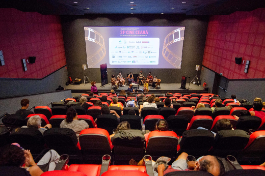 Imagem: Sala de cinema com poltronas vermelhas e pessoas sentadas de costas viradas para tela de cinema ao fundo.