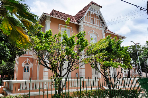 Imagem: Casa de dois andares, em estilo alemão, na cor rosa, com árvores na frente.