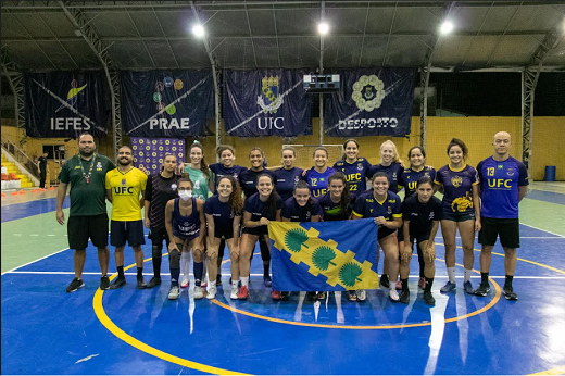 Imagem: Equipe de futsal feminino, atletas em posição com auxiliares técnicos dentro de um ginásio com piso azul