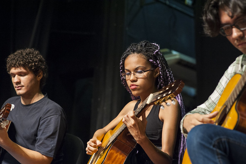 Imagem: foto de três jovens tocando violão. Da esquerda para a direita, um rapaz negro de cabelos curtos; uma moça negra de cabelo trançado e um rapaz branco.