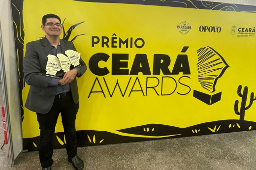 Imagem: professor Abraão Saraiva segura os três trofeus recebidos em frente a um banner amarelo com os dizeres "prêmio Ceará Awards"