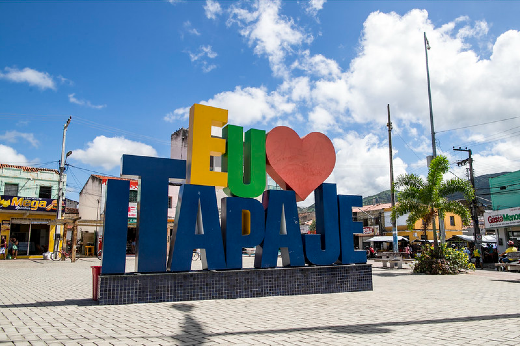 Imagem: Letreiro com a frase "Eu amo Itapajé", em letras coloridas, localizado no meio da praça. Céu azul e fundo com comércio.