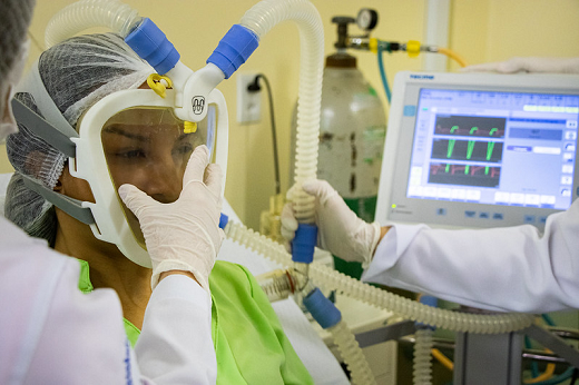 Imagem: Paciente usando máscara respiratório que cobra o rosto inteiro; a mão de uma enfermeira toca a parte da frente da máscara, e outra toca um tubo deligação