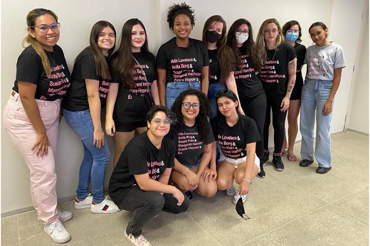 Várias jovens posam para a foto. Eles estão com uma blusa preta com uma lista de nomes de mulheres na cor lilás