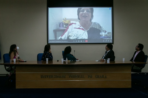 Imagem de mulher com cabelo preto projetada em tela de auditório e cinco pessoas sentadas na mesa