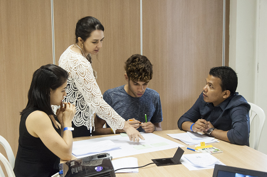 Imagem: Quatro estudantes (uma de  pé e três sentados) discutem com papéis sobre uma mesa, durante Feira de Empreendedorismo da UFC (Foto: Viktor Braga/ UFC Informa)