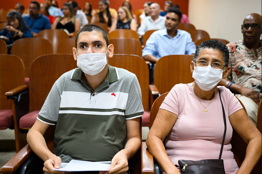 Imagem: Homem de blusa cinza sentado em ambiente de auditório ao lado de uma senhora com blusa rosa