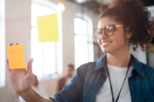 Imagem: Jovem estudante organiza post-its coloridos em janela de vidro. É uma mulher negra, de óculos, camiseta branca, camisa jeans aberta e crachá 