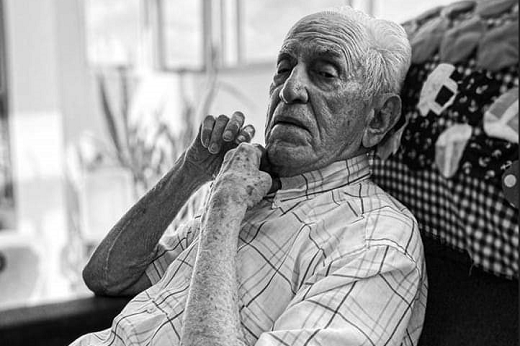Foto em preto e branco de B de Paiva, um senhor idos, de cabelos brancos, que veste camiseta de botão e está sentado