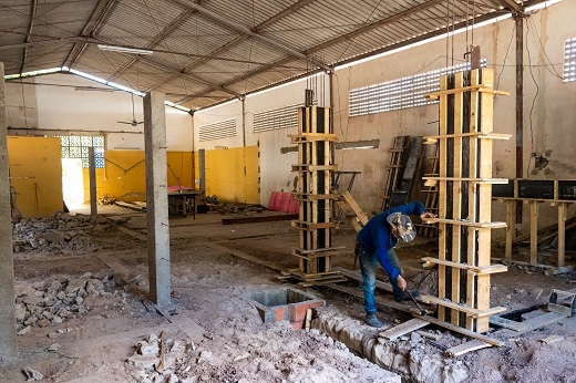 Imagem: Trabalhador agachado, vestido de azul e usando chapéu, trabalha em canteiro de obras (ambiente interno) na UFC. (Foto: Ribamar Neto)