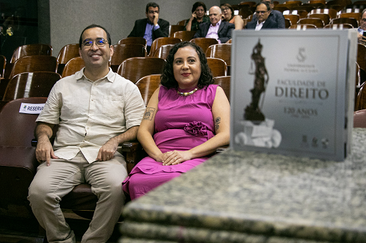 Imagem: Organizadores do livro, os jornalistas Marco Fukuda e Cristiane Pimentel posam sentados no anfiteatro com o livro comemorativo em primeiro plano. (Foto: Ribamar Neto/ UFC Informa)